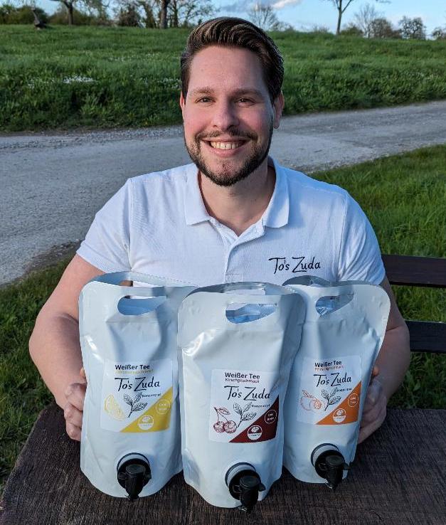 Dieses Bild zeigt den Gründer von To's Zuda, Tobias, der auf einer Bank an einem Feldweg sitzt. Auf dem Tisch vor ihm stehen drei Verpackungen mit To's Zuda Sirup, mit den Geschmacksrichtungen Zitrone, Kirsche und Pfirsich.